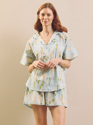 A woman modelling a light blue short-sleeve pyjama set with a light blue and yellow floral print, consisting of a button-up top and matching shorts, against a solid beige background.