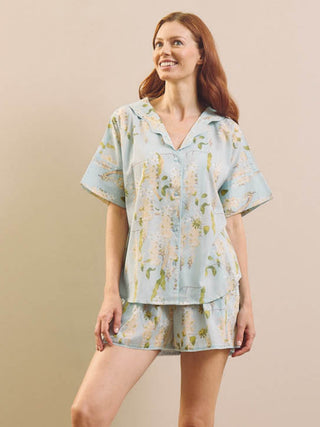 model wears a pair of short pyjamas in a light fabric in pale blue with white wisteria against a beige wall. 