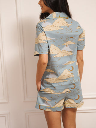 Model wearing women’s cotton short pyjama set in light blue with cloudy skies print, featuring short-sleeve button-front shirt with revere collar and matching shorts with pockets. Back detail