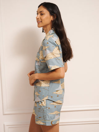 Model wearing women’s cotton short pyjama set in light blue with cloudy skies print, featuring short-sleeve button-front shirt with revere collar and matching shorts with pockets.