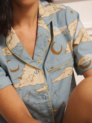 Model wearing women’s cotton short pyjama set in light blue with cloudy skies print, featuring short-sleeve button-front shirt with revere collar and matching shorts with pockets.