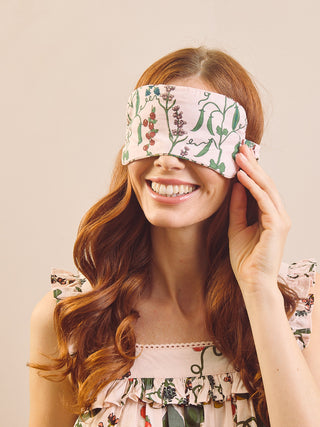 A smiling woman with long wavy auburn hair wears a light pink floral blindfold and a matching ruffled top against a plain beige background.