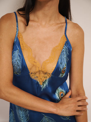 Woman wearing a blue  and gold night dress slip with peacock feather pattern against a white wall.