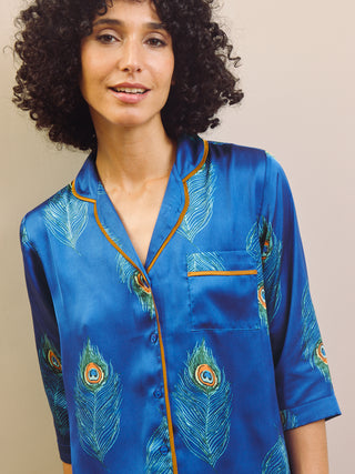 Woman wearing a blue satin pajama top with peacock feather patterns against a beige background