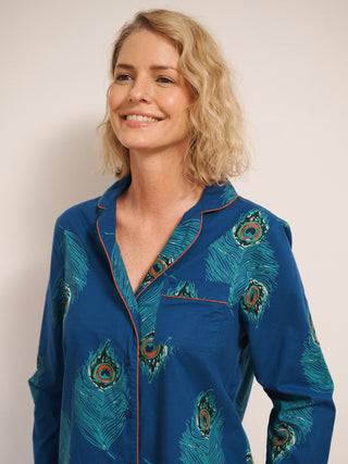 Woman wearing a blue pyjama shirt with peacock feather patterns on a plain background