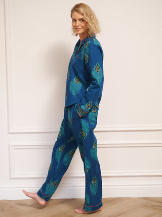 Woman wearing a blue pyjama set with peacock patterns against a white wall.