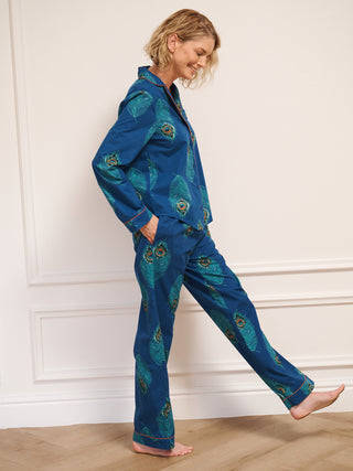 Woman wearing a blue pyjama set with peacock patterns against a white wall.