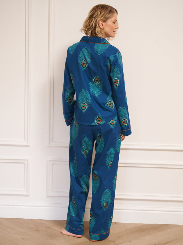 Woman wearing a blue pyjama set with peacock feather pattern against a white wall.