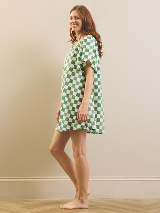A woman modeling a short-sleeve nightgown with a green and white checkered pattern, standing barefoot against a beige wall and light wood floor.
