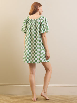 A woman stands barefoot in a studio wearing a short-sleeve green and white checkered nightgown, photographed from the back against a plain beige wall and light wood floor.
