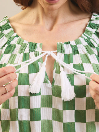 Close-up of a woman wearing a green and white checkered top with white tassels at the neckline, holding the tie strings with both hands.