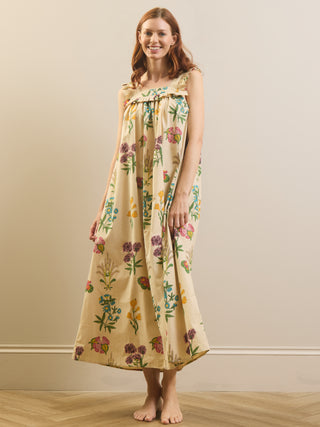 A woman stands barefoot in a studio wearing a long cream-colored nightgown with a colorful floral and botanical print. The dress features ruffled shoulder straps and a gathered neckline.
