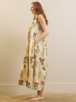 A woman wearing a sleeveless cream-colored maxi nightdress with a colorful floral and botanical print, standing barefoot in a room with a beige wall and light wood floor.
