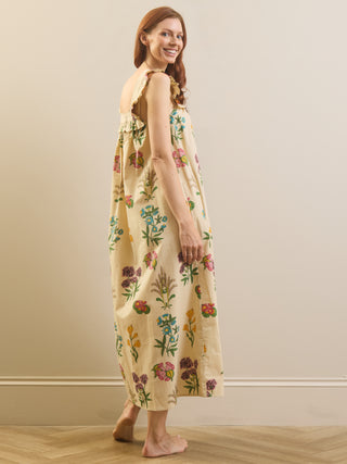 A woman wearing a sleeveless cream-colored nightdress with a colourful floral print, standing barefoot in a room with a beige wall and light wood floor.