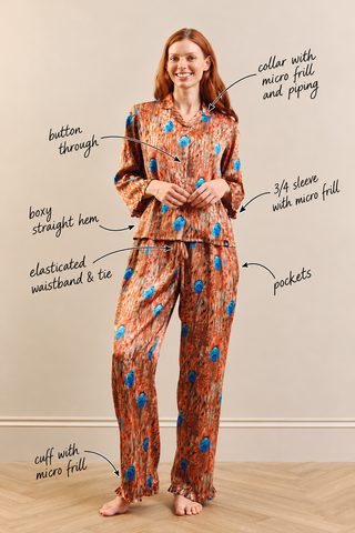 A woman modeling a two-piece pajama set with hand-drawn callout lines and text labels pointing to features like the collar, button, sleeves, hem, waistband, cuffs, and pockets. The pajamas have a warm brown and orange abstract pattern with bright blue circular accents.