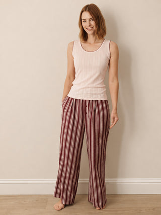 A woman stands barefoot against a plain beige wall wearing a light pink ribbed tank top and burgundy-and-cream vertically striped wide-leg pants.
