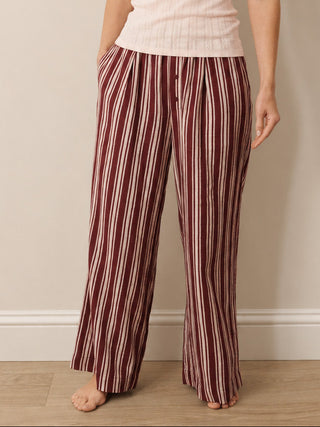 A woman stands barefoot against a plain beige wall wearing a light pink ribbed tank top and burgundy-and-cream vertically striped wide-leg pants.