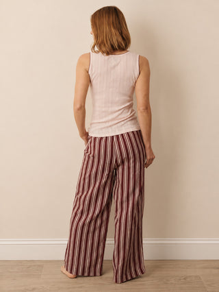 A woman viewed from behind wearing a light pink ribbed sleeveless top and wide-leg trousers with vertical burgundy and cream stripes, standing against a beige wall.