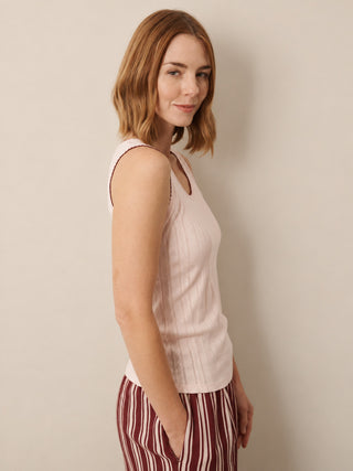 A woman with shoulder-length auburn hair is shown in profile wearing a light pink ribbed knit sleeveless top and burgundy-and-cream vertically striped trousers, standing against a plain beige wall.