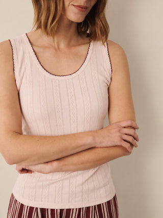 A woman wearing a light pink ribbed knit tank top with dark red scalloped trim along the scoop neckline, paired with a striped skirt, against a neutral beige background.