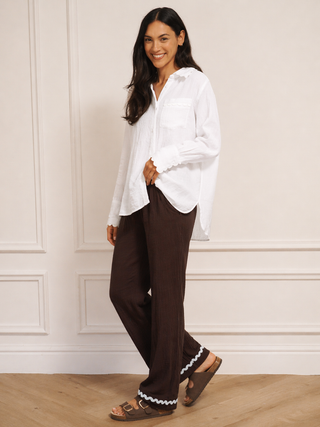 Woman wearing a white blouse and brown pants with white trim against a beige wall.