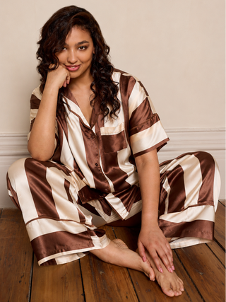 Woman wearing a brown and white striped satin pajama set sitting on a wooden floor.
