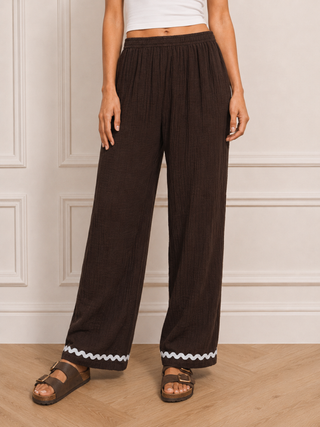 A person wearing dark brown wide-leg pleated pants with a white zigzag trim at the hem, paired with brown sandals and a white cropped top, standing in front of a cream paneled wall.