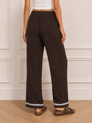 A person wearing dark brown wide-leg pleated pants with a white zigzag trim at the hem, paired with a white crop top and brown slide sandals, standing against a white paneled wall.