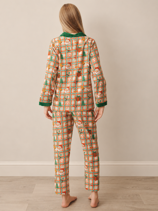 A woman stands with her back to the camera wearing a matching long-sleeve pyjama set with a festive holiday print of Santa Claus and Christmas trees on a beige background with green and orange accents.