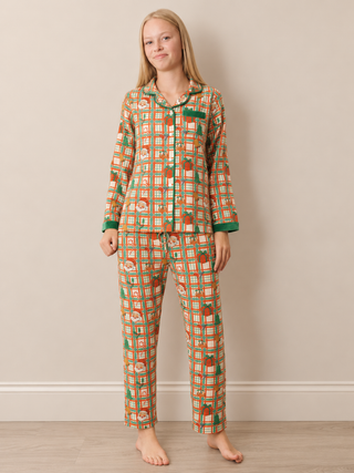 A young girl stands barefoot against a beige wall wearing a long-sleeve button-up pyjama set with an orange, green, and white geometric pattern.