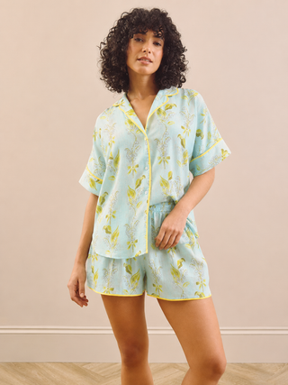 A woman modelling a two-piece pyjama set consisting of a short-sleeve button-front shirt and matching shorts in a light pastel blue with a yellow floral print and yellow trim, set against a solid beige wall.
