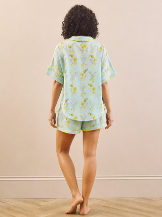 A woman stands with her back to the camera wearing a light mint green short-sleeve pajama shirt and matching shorts with a yellow floral print, set against a beige wall and light wood herringbone floor.