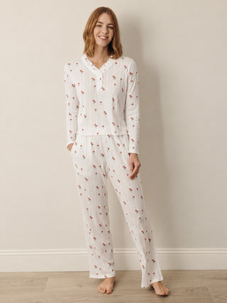 A woman modeling a two-piece white long-sleeve pyjama set with a small red floral print, standing against a neutral beige wall and light wood floor.