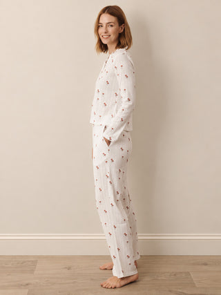 A woman modelling a two-piece white pyjama set with a small red floral pattern, standing barefoot against a beige wall and light wood floor.