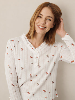 A smiling woman with light brown hair wears a white long-sleeve pyjama top with a small red mushroom print and vertical lace-like stripes, posed against a plain beige background.