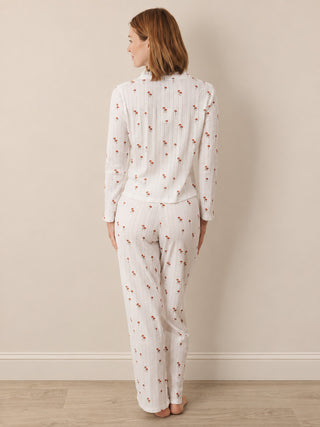 A woman stands with her back to the camera wearing a matching white long-sleeve top and wide-leg pant pyjama set, both featuring a small red floral print, in a neutral indoor setting.