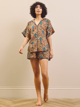 A woman modelling a matching short-sleeve button-front top and shorts pyjama set with an abstract orange, brown, and teal pattern, standing barefoot against a beige wall.