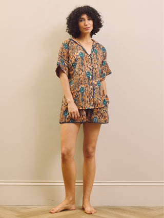 A woman modelling a matching short-sleeve button-front top and shorts pyjama set with an abstract orange, brown, and teal pattern, standing barefoot against a beige wall.