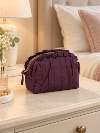 A deep purple pleated cosmetic pouch with gold-tone hardware, resting on a cream-colored bedside table next to a lamp and flowers.