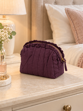 A deep purple quilted cosmetic pouch with gold-tone zippers, resting on a cream marble bedside table in a warm, softly lit bedroom setting.
