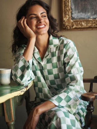 A woman smiling while wearing a green and white checkered pyjama set, sitting in a rustic indoor setting next to a table.