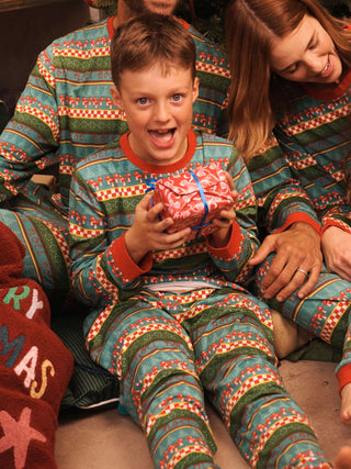 A family wearing matching Christmas pyjamas sits together indoors, with a young boy in the center holding a wrapped gift. The pajamas have a festive multicolor pattern with red, green, blue, and white holiday motifs.