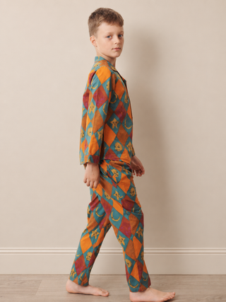 A young boy wearing a two-piece long-sleeve pyjama set with a colorful harlequin diamond pattern, walking barefoot against a neutral beige wall.