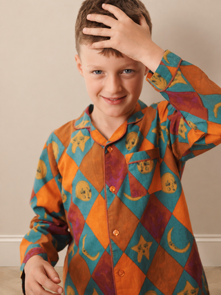 A young boy wearing a long-sleeve button-up pyjama shirt with a colourful harlequin diamond pattern in orange, teal, and purple, featuring skull and crescent moon motifs. He is smiling and touching his hair against a plain beige wall background.