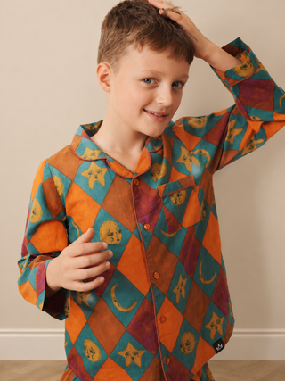 A young boy wearing a long-sleeve button-up pyjama shirt with a colourful harlequin diamond pattern of orange, teal, purple, and brown, featuring celestial motifs like stars, moons, and skulls.