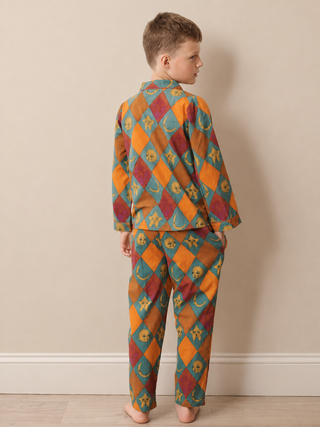 A child standing with their back to the camera, wearing a matching two-piece pyjama set with a colorful harlequin diamond pattern in orange, teal, and burgundy with yellow crescent moon details, set against a plain beige wall.