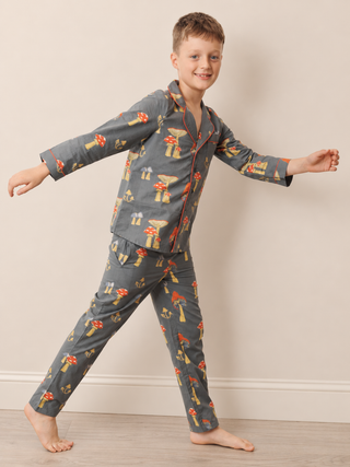 A young boy wearing a gray long-sleeve pyjama set with a red and yellow mushroom print, posing barefoot against a neutral beige wall.