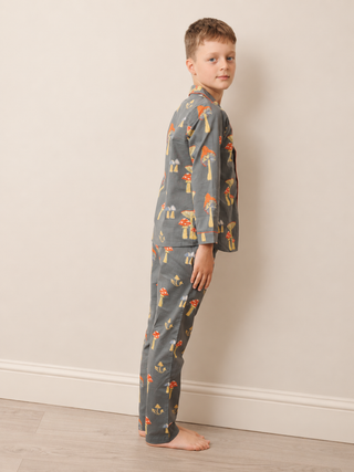 A young boy stands barefoot in profile against a plain beige wall, wearing a two-piece pyjama set with a gray base and a colorful mushroom print.