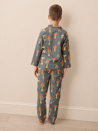 A child standing with their back to the camera wearing a two-piece pyjama set in dark gray with a colorful mushroom print, set against a plain beige wall and light wood floor.