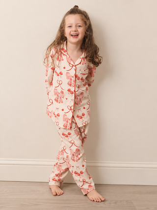A young girl with long light brown hair stands barefoot against a beige wall, wearing a matching long-sleeve pyjama set with a red bow and ribbon pattern on a cream background.